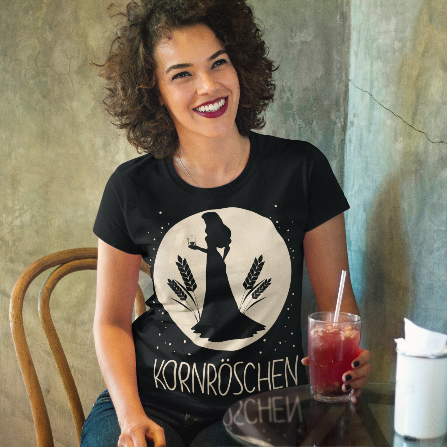 Eine Frau trägt das Kornröschen Shirt an der Bar und trinkt einen roten Cocktail am Tisch